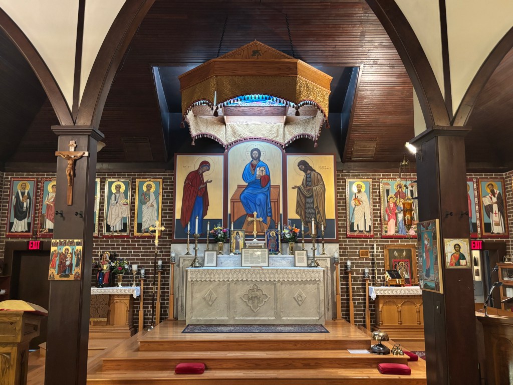 Interior view of Saint Mark's Parish featuring an altar adorned with religious icons, candles, and a decorative canopy. The walls are decorated with framed icons of saints.
